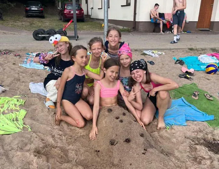 Grupa uczestniczek w strojach kąpielowych pozuje na piaszczystej plaży wśród ręczników