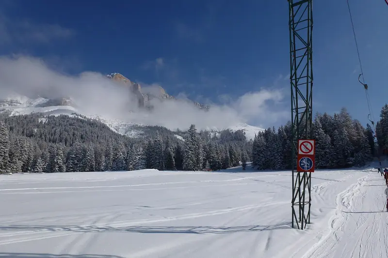 Wyratrakowana trasa narciarska w Carezza w słoneczny dzień, podpora wyciągu orczykowego z boku, w tle dolomickie ściany we mgle