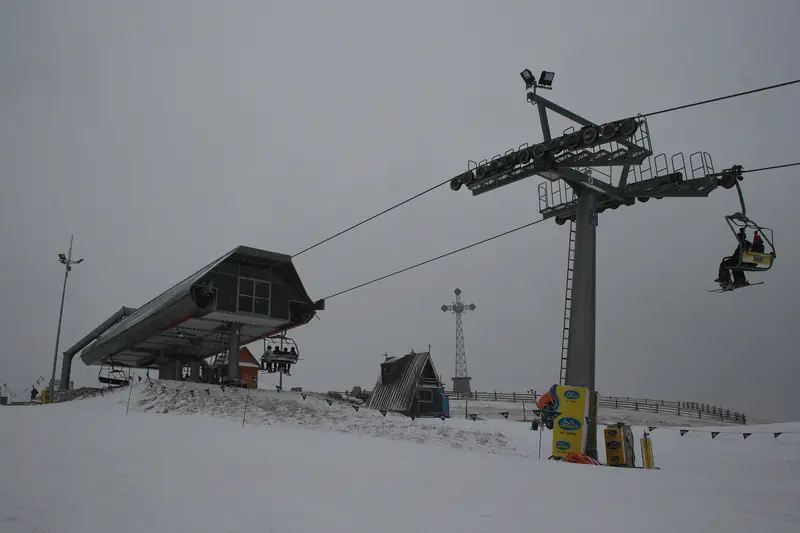 The upper terminal of the Grapa-Litwinka chairlift in Czarna Góra, skiers on the chairs above a snowy slope, the Millennium Cross behind