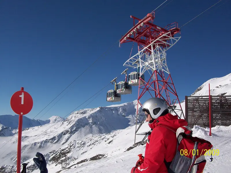 Podpora kolei linowej Ankogelbahn nad ośnieżonym stokiem w Mallnitz, gondole nad głębokim śniegiem, narciarz i słupek trasy pod błękitnym niebem