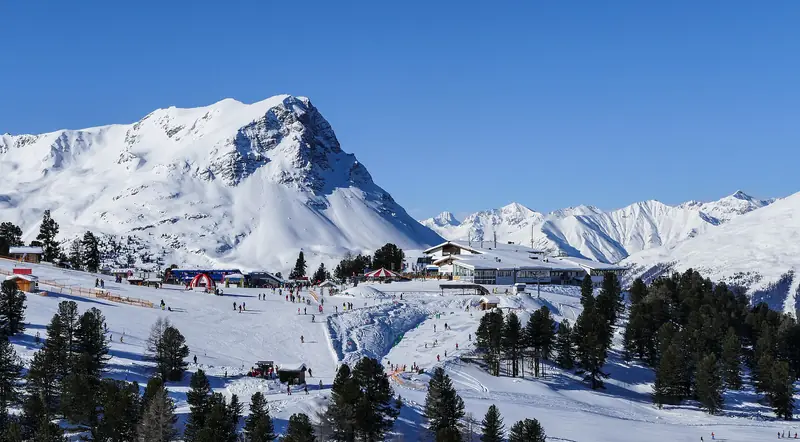 Ośrodek Bergkastel w Nauders: skalisty szczyt Bergkastelspitze nad stacją dolną i szerokimi trasami, narciarze na śniegu w słońcu