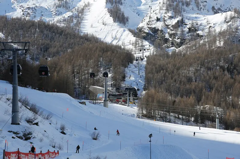 A gondola above a snowy Saas-Fee piste among the larches, skiers below