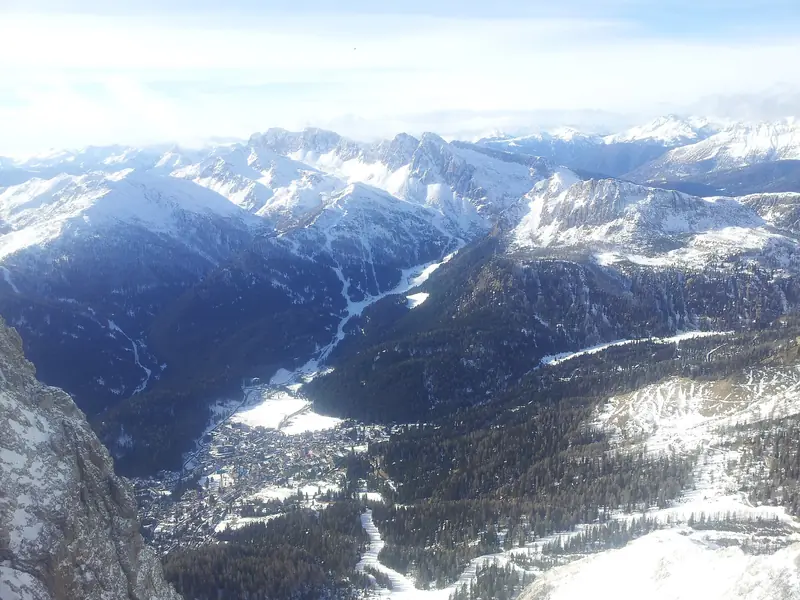 Widok z gondoli na San Martino di Castrozza w dolinie, trasy narciarskie schodzą lasem do miasteczka, wokół ośnieżone szczyty Dolomitów