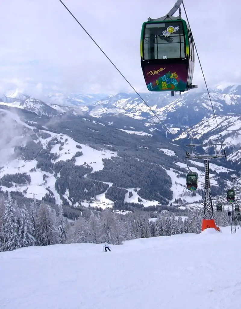 A red gondola above a snowy Wagrain piste, a panorama of the snowy Salzburg Alps, a skier below