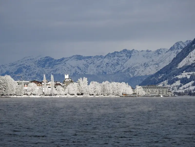 Zell am See zimą nad jeziorem Zeller See, oszronione drzewa i zabudowa nad brzegiem, w tle ośnieżone szczyty Hohe Tauern
